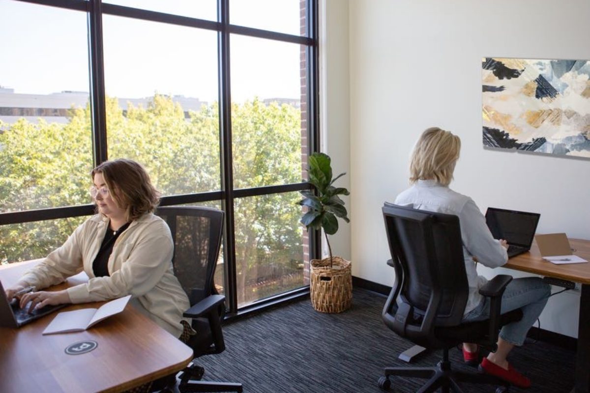 Two people working at sit/stand desks in ergonomic chairs at The District Coworking and Flex Office in Eugene, Oregon, near a large window.