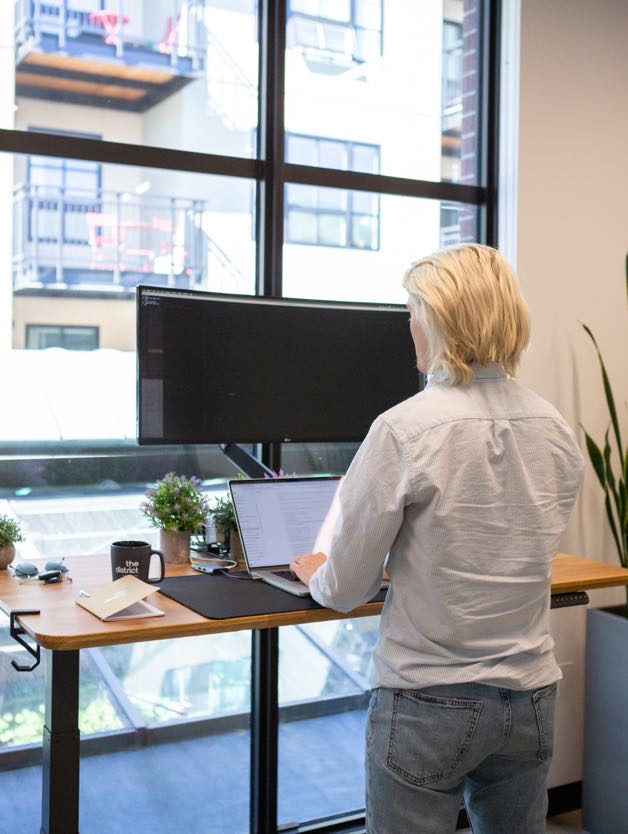 Person working at a dedicated desk in front of a large bright window at The District Coworking and Flex Office in Eugene, Oregon, in a modern and professional workspace.